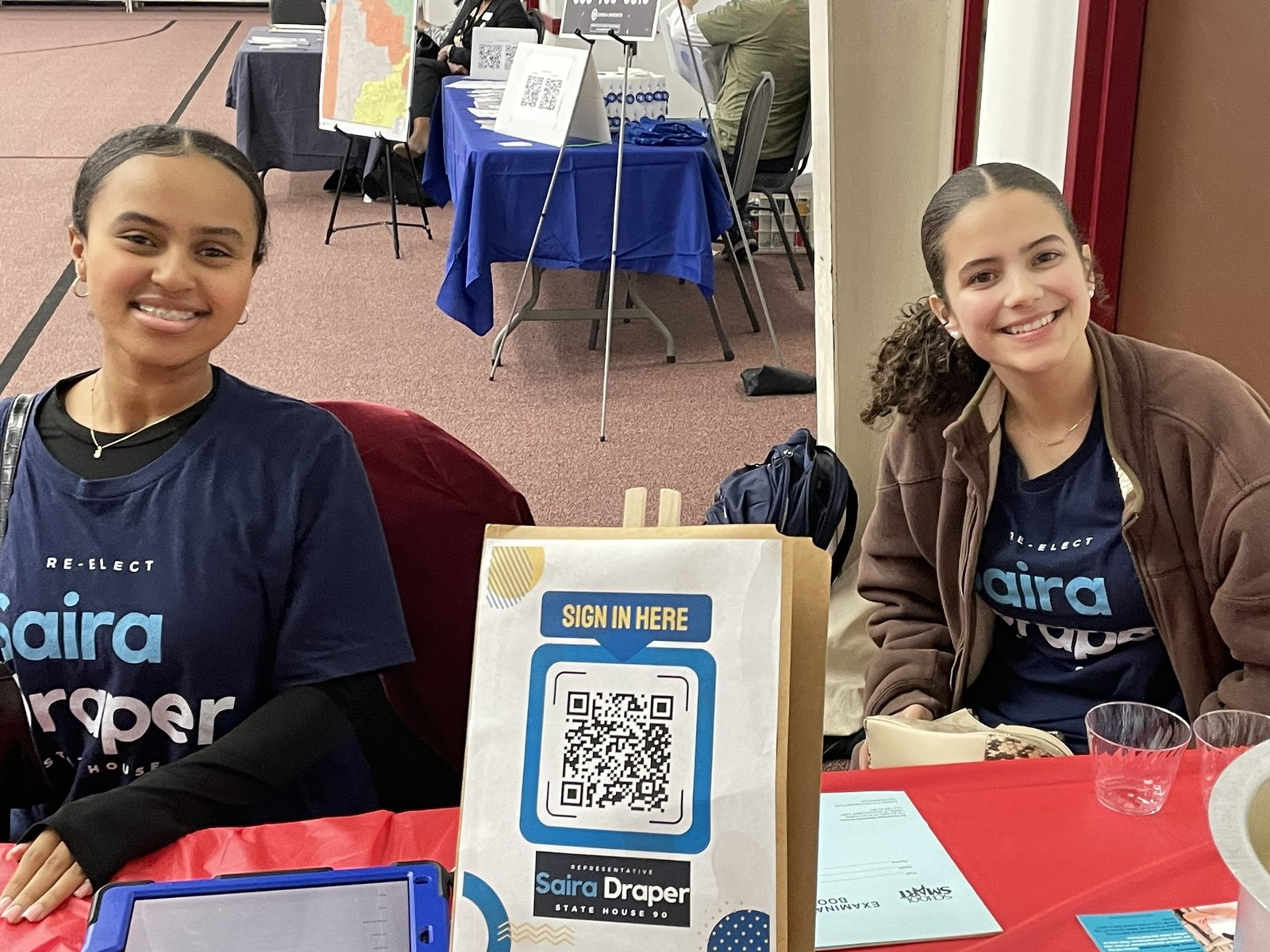Two campaign volunteers in Re-Elect Saira Draper t-shirts smiling at a community event sign-in table